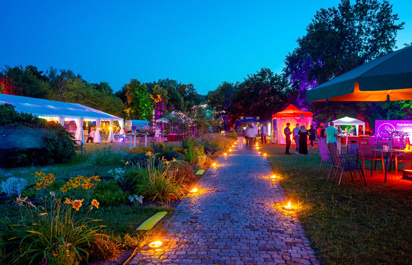 Ein Foto von einem schön beleuchteten Parkgarten mit Gästen und vielen Fest-Zelten
